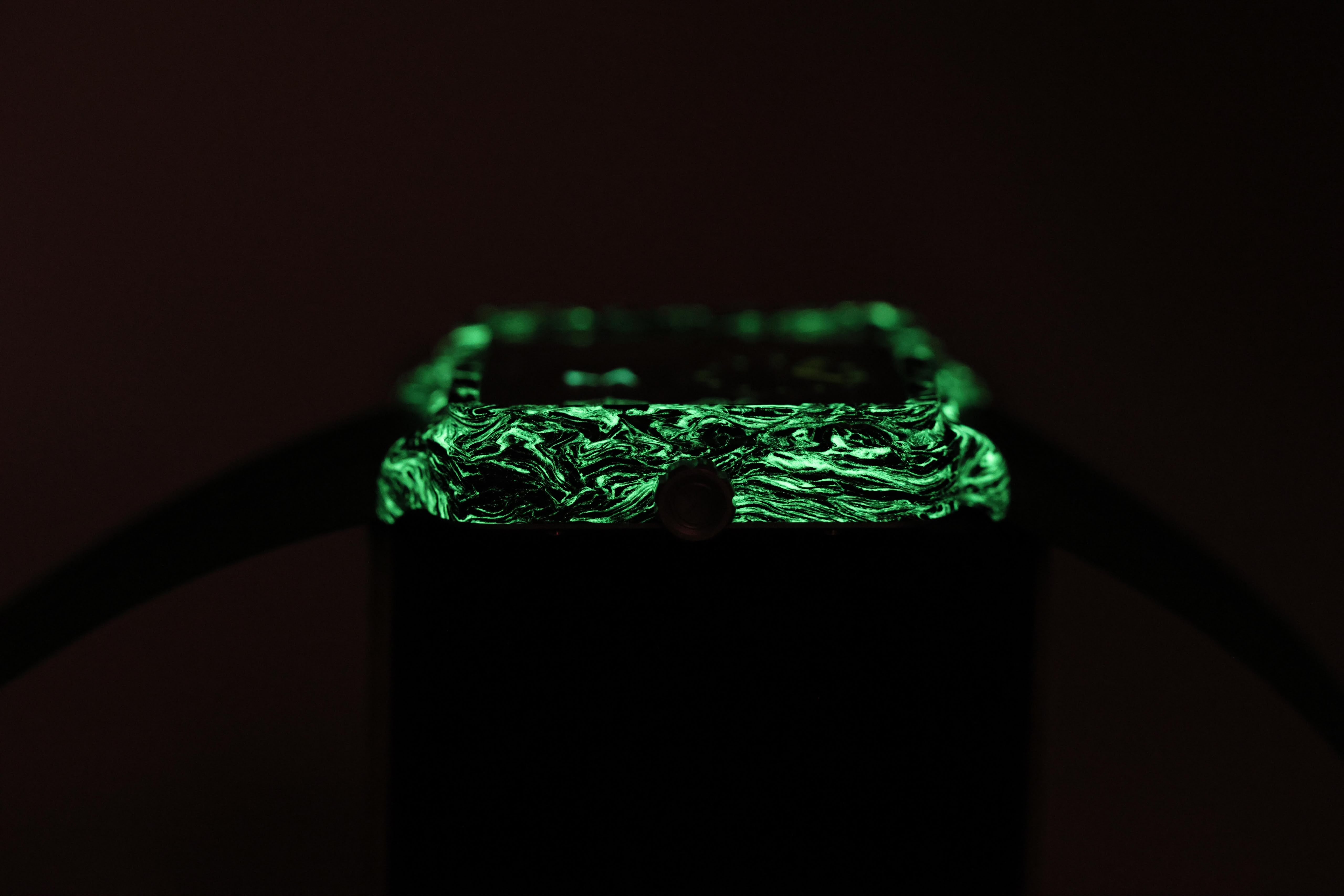 A close-up of a black rectangular watch with a glowing green marbled or textured top surface, photographed in very dark lighting against a black background.