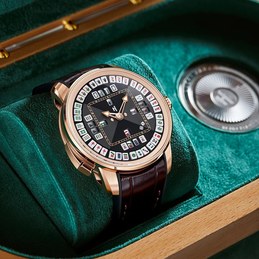 A luxury watch with a rose gold case and a dark brown leather strap, resting on a plush green velvet cushion inside a wooden presentation box. The watch face features a black background with a circular array of colorful, patterned tiles and gold hands.