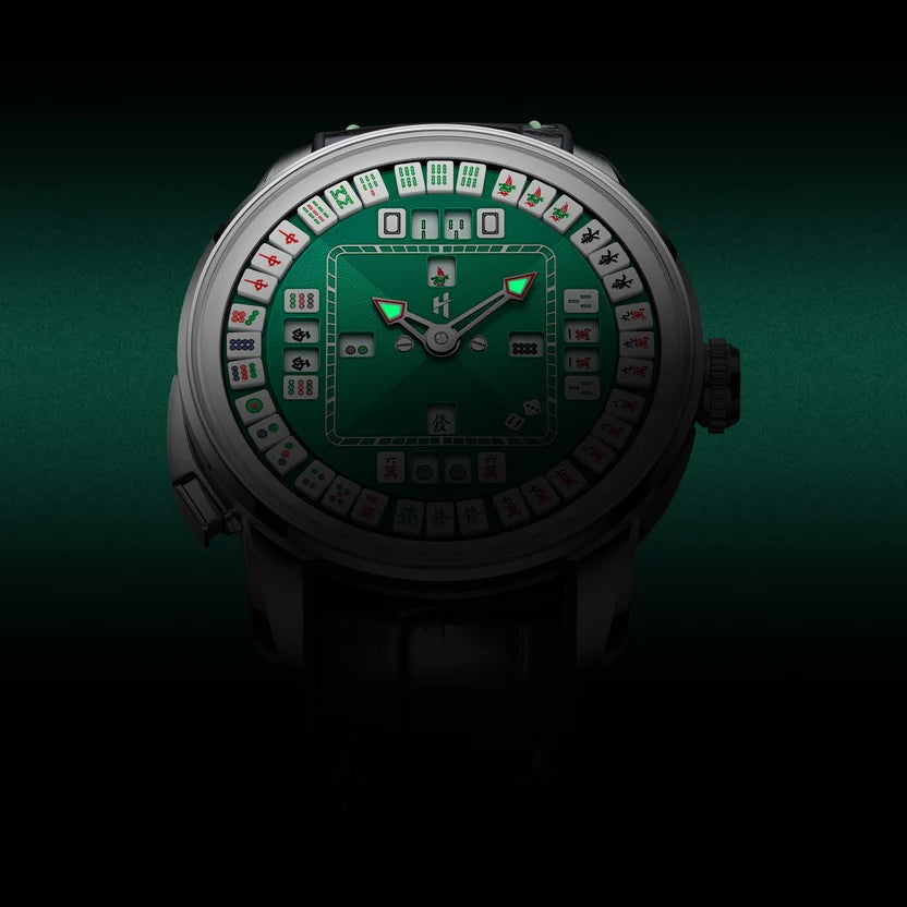 A luxury watch with a dark green mahjong-themed dial, featuring illuminated green hands and white, red, and black tile graphics around the inner ring, shown against a dark green and black background.