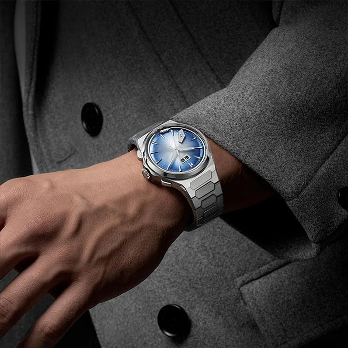A silver-toned metal watch with a blue sunburst dial and matching blue bezel, worn on a person's wrist. The person is wearing a dark gray textured blazer.