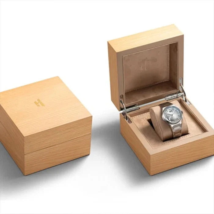 A light natural wood watch presentation box shown closed on the left and open on the right, with a silver-toned watch displayed inside on a cushion. The open box has a transparent top panel and a small gold monogram logo on the lid.