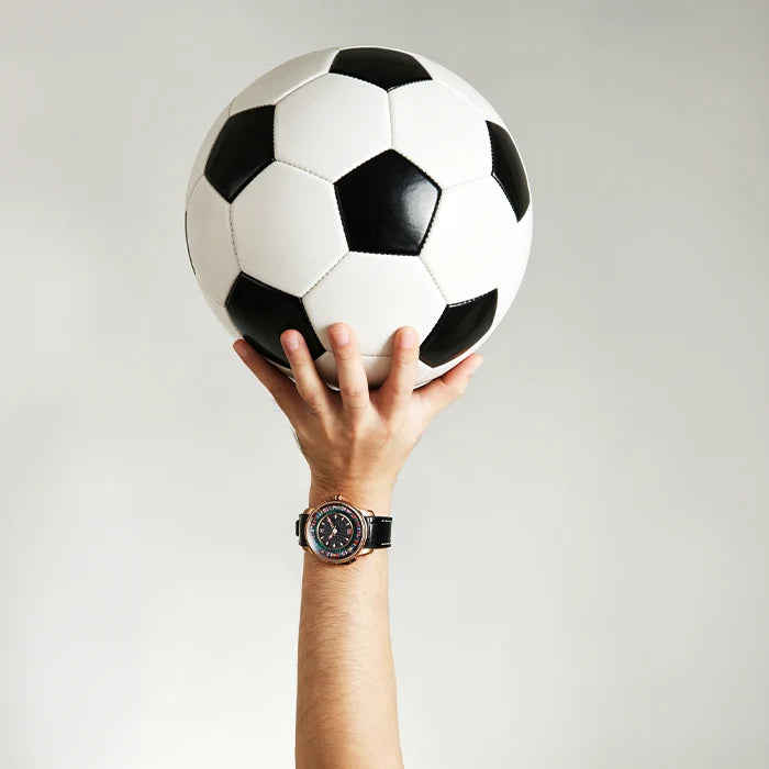 A hand holding a classic black and white soccer ball, with a wristwatch visible on the wrist, set against a plain light gray background.