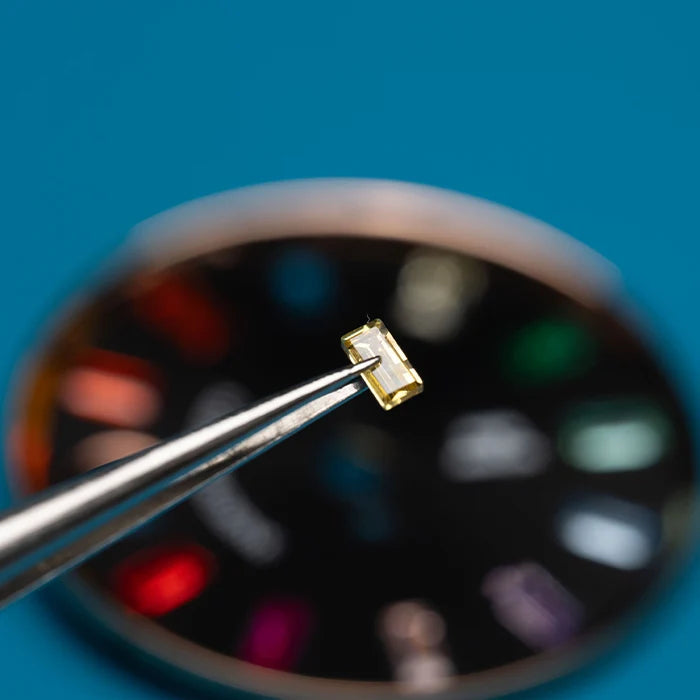A small rectangular yellow gemstone held by metal tweezers in sharp focus against a blurred blue background with a circular color wheel.
