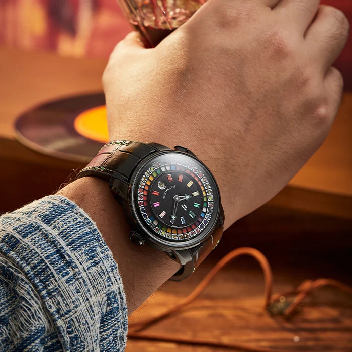 A close-up lifestyle shot of a person's wrist wearing a black smartwatch with a colorful dial and a dark leather strap, set against a warm, blurred background with a vinyl record and an orange cable.
