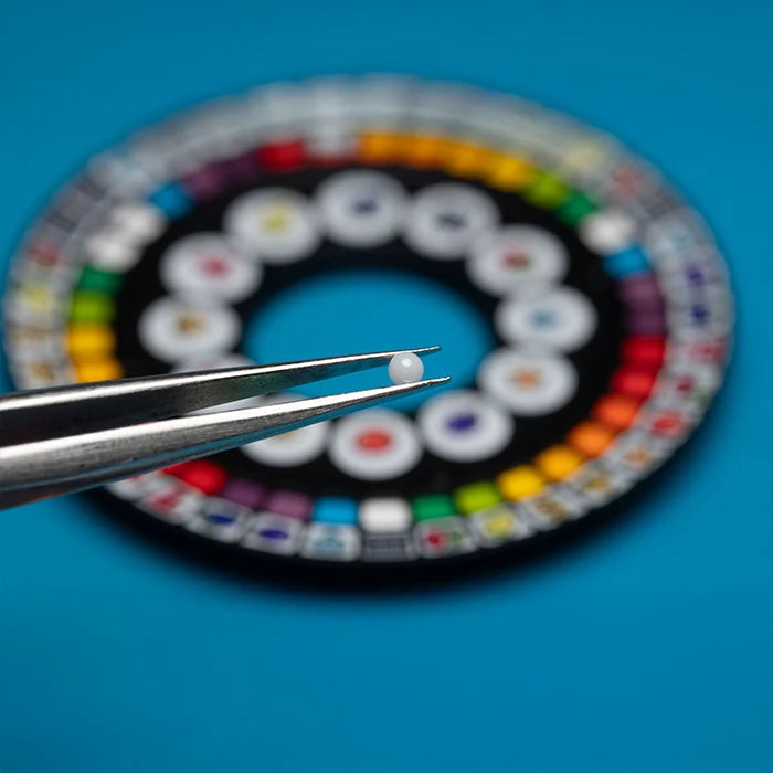 A tiny round white gemstone or bead held in the jaws of metal tweezers, with a blurred multicolor circular color wheel on a bright blue background.