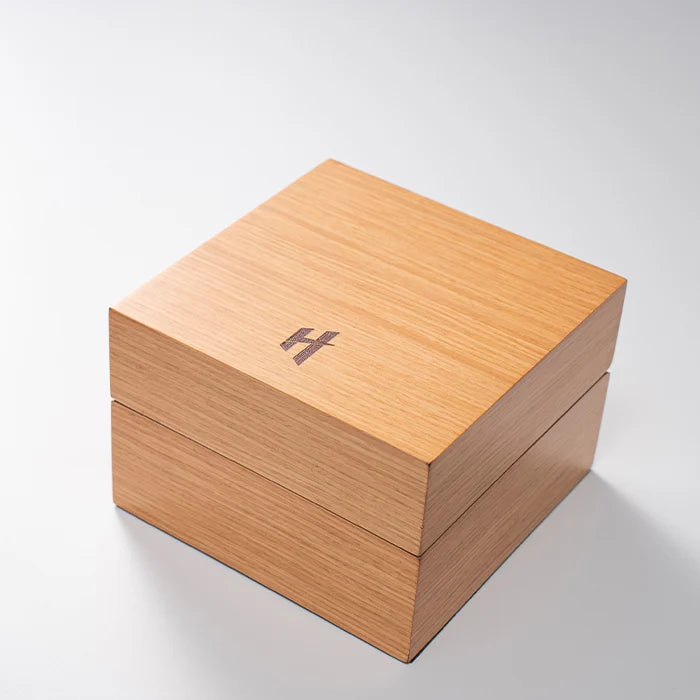 A square light wood presentation box with a hinged lid, featuring an engraved dark logo on the top surface, set against a clean white background.