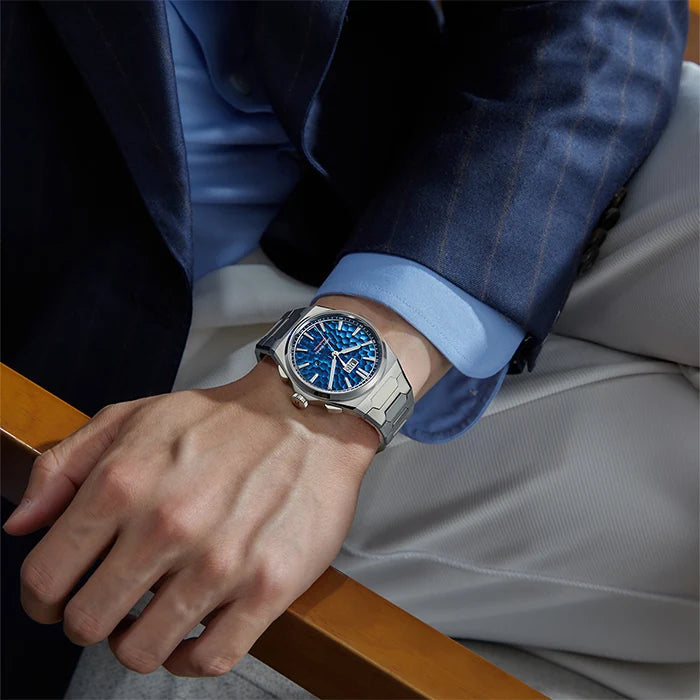 Person wearing a watch with a blue face, dressed in a suit, on a wooden surface.