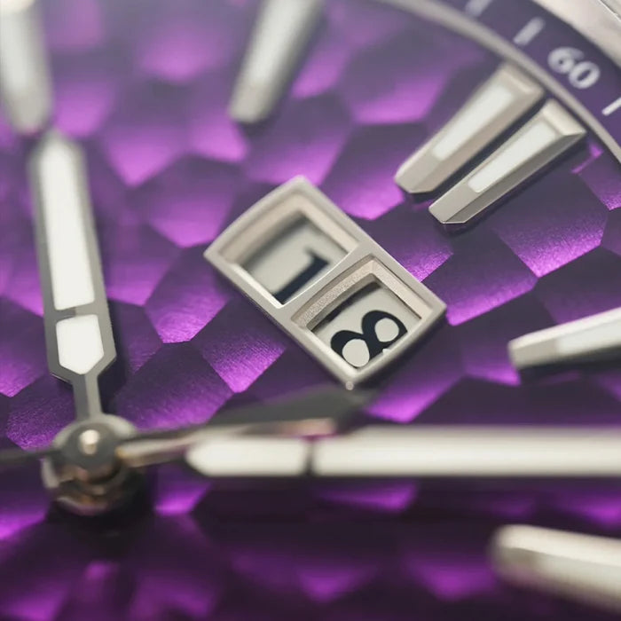 Close-up of a purple textured dial watch face with silver-tone hands, hour markers, and a date window showing 18.
