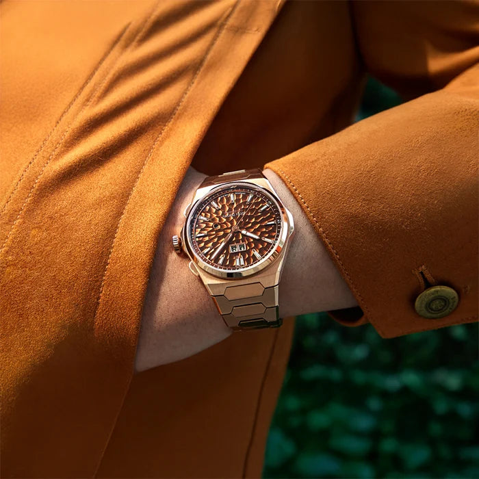 Close-up of a person wearing a gold watch with a detailed face, against an orange coat background.