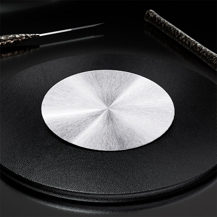 A round brushed silver metal coaster or trivet pad placed on top of a black textured circular mat, with dark background and partial decorative objects at the top corners.