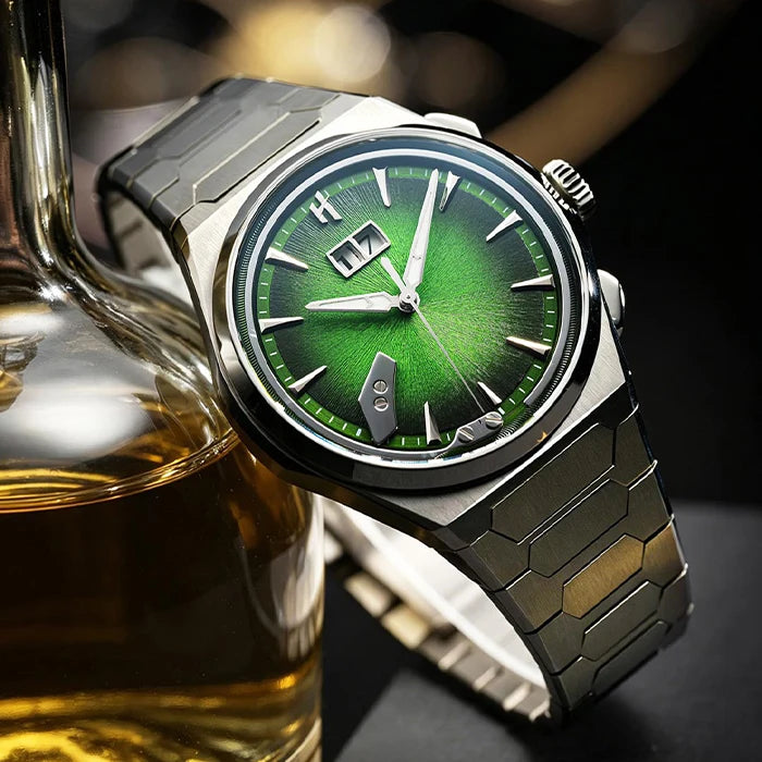 A titanium  wristwatch with a green sunburst dial, silver-tone hour markers, and a matching metal link bracelet, styled next to a glass decanter of amber liquid on a dark surface.