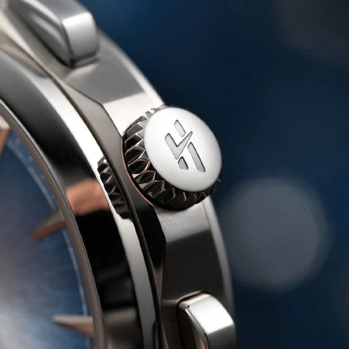 Close-up of a stainless steel watch case showing the crown and pushers, with a blue dial partially visible and a polished metallic finish against a blurred dark blue background.