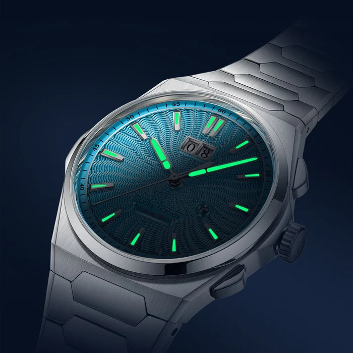A close-up of a silver-toned stainless steel watch with a textured blue-green dial and bright green luminescent hour markers and hands, shown against a dark navy background.