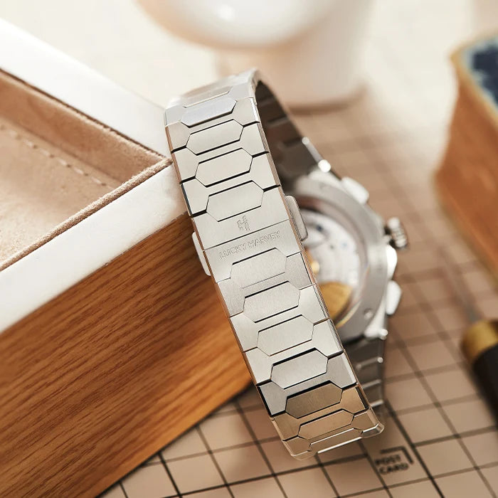 A close-up of a silver stainless steel link bracelet watch resting against a wooden box on a checkered surface, with the watch case partially visible behind it.