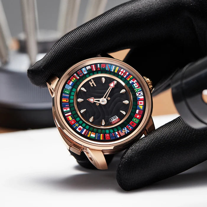 A close-up of a luxury watch with a rose gold case and a black dial decorated with colorful international flags, held by a hand wearing a black glove.