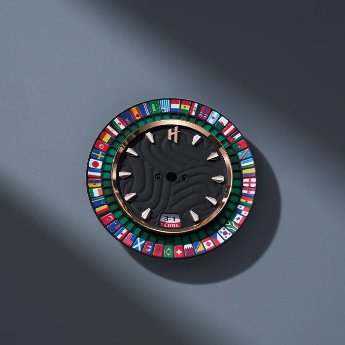 A round wall clock with a black face, gold-tone rim, and a colorful outer ring featuring many national flags, mounted on a dark gray wall with a soft shadow.