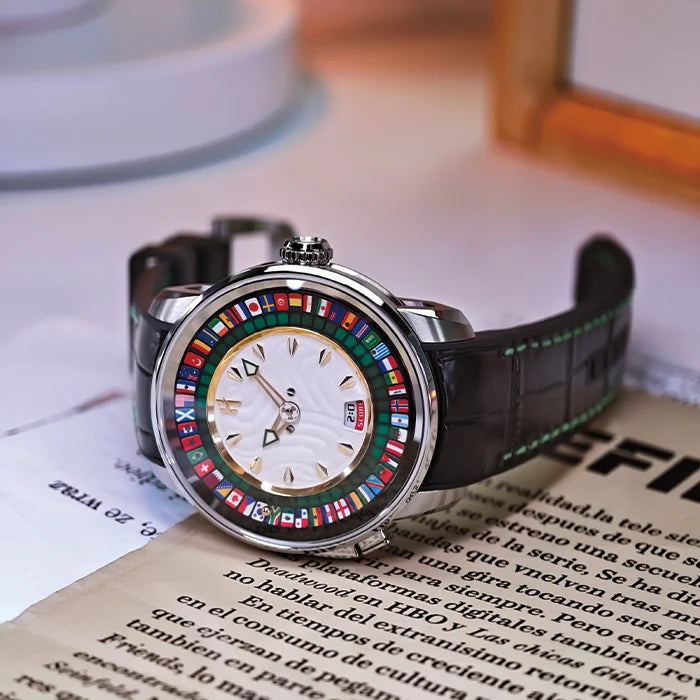 A luxury watch with a silver-toned case, a mother-of-pearl dial, and a rotating ring of colorful national flags. The watch has a black alligator leather strap with green stitching and is resting on an open book with Spanish text.
