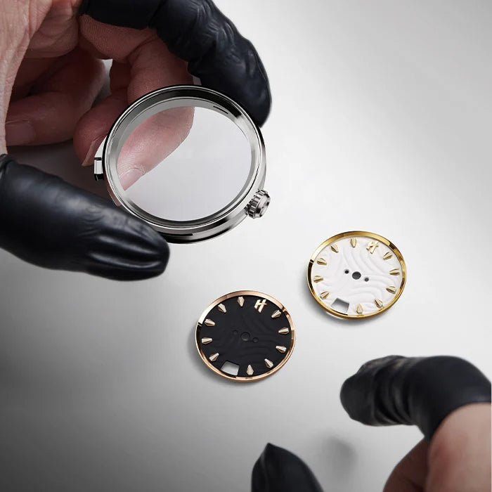 Person wearing black gloves examining watch dials with a magnifying glass on a light gray background